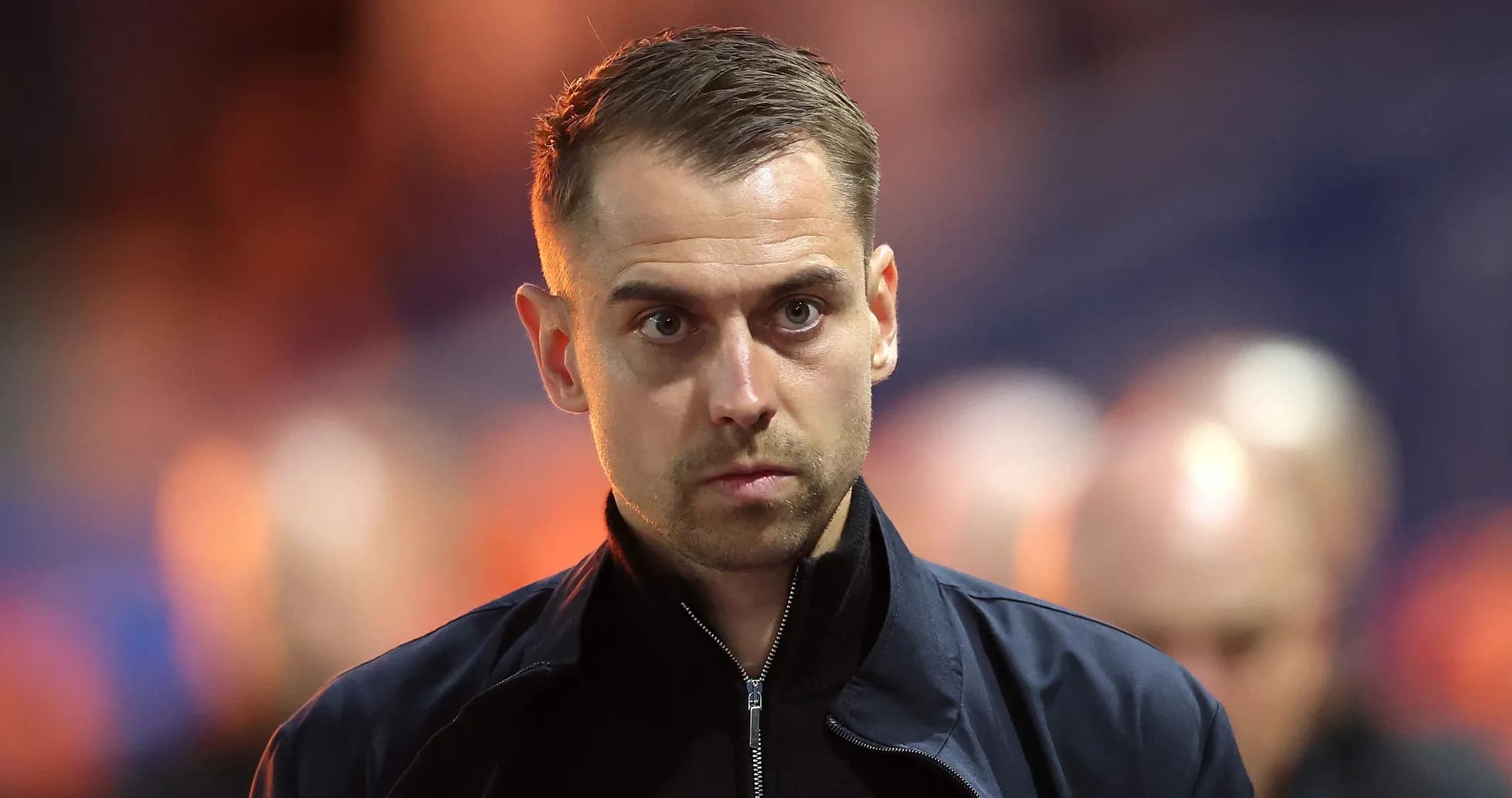Man in dark jacket walking on stadium sidelines